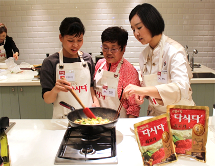 다시다쇠고기 제품이 놓인 조리대 앞에서 요리사와 다문화가정의 외국인 며느리와 시어머니가 다정히 서서 프라이팬에 놓인 음식을 요리하고 있다.