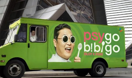 PSYgo bibigo라는 문구와 가수 싸이의 얼굴이 그려진 녹색 트럭 운전석에 미소띈 얼굴의 싸이가 탑승해 있다. 트럭 뒤로는 건물이 보인다.
