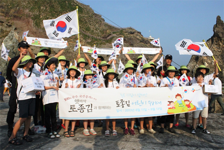 햇바삭 토종김과 함께하는 어린이 수비대 10명과 보호자 10명이 독도에서 태극기를 흔들며 기념사진을 찍고있다.